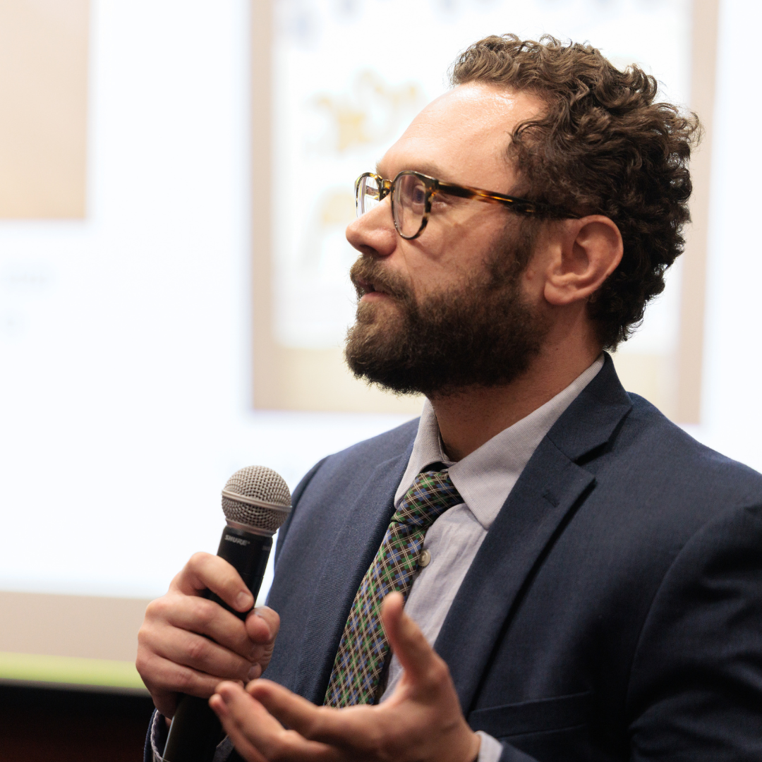 A young man with curly brown hair, a full brown beard and glasses is wearing a navy suit coat, white shirt and tie. He is holding a microphone in his right hand and gesturing with his left while he is speaking.